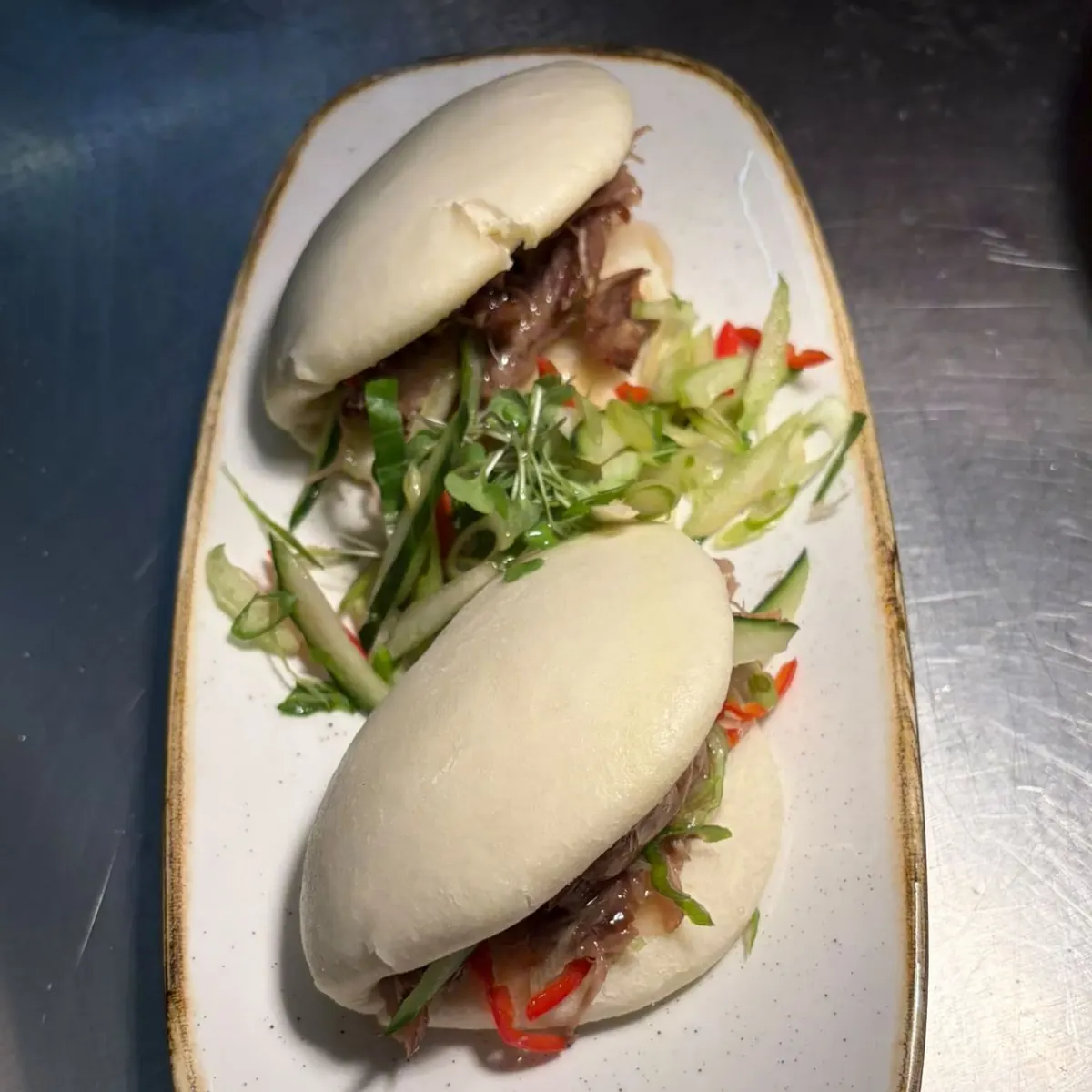 Two steamed bao buns filled with shredded duck, with a cucumber, spring onion and chilli salad