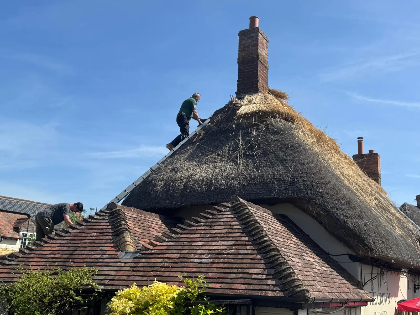 Two thatchers working on the roof of The Crown, laying fresh straw along the ridge beside the chimney