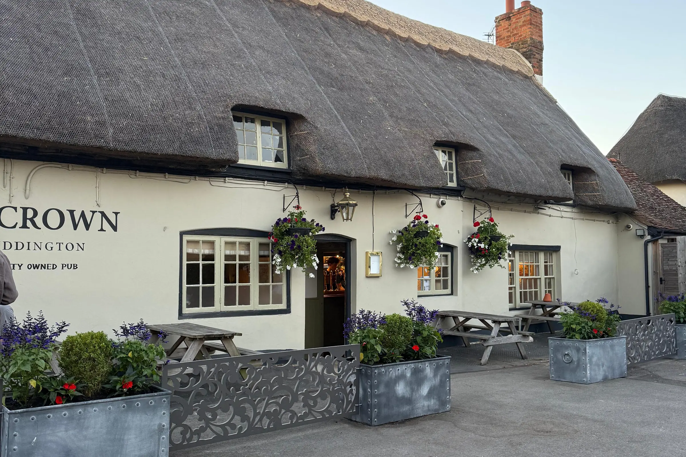 The thatched front of The Crown at dusk, hanging baskets in flower and the door open onto the bar
