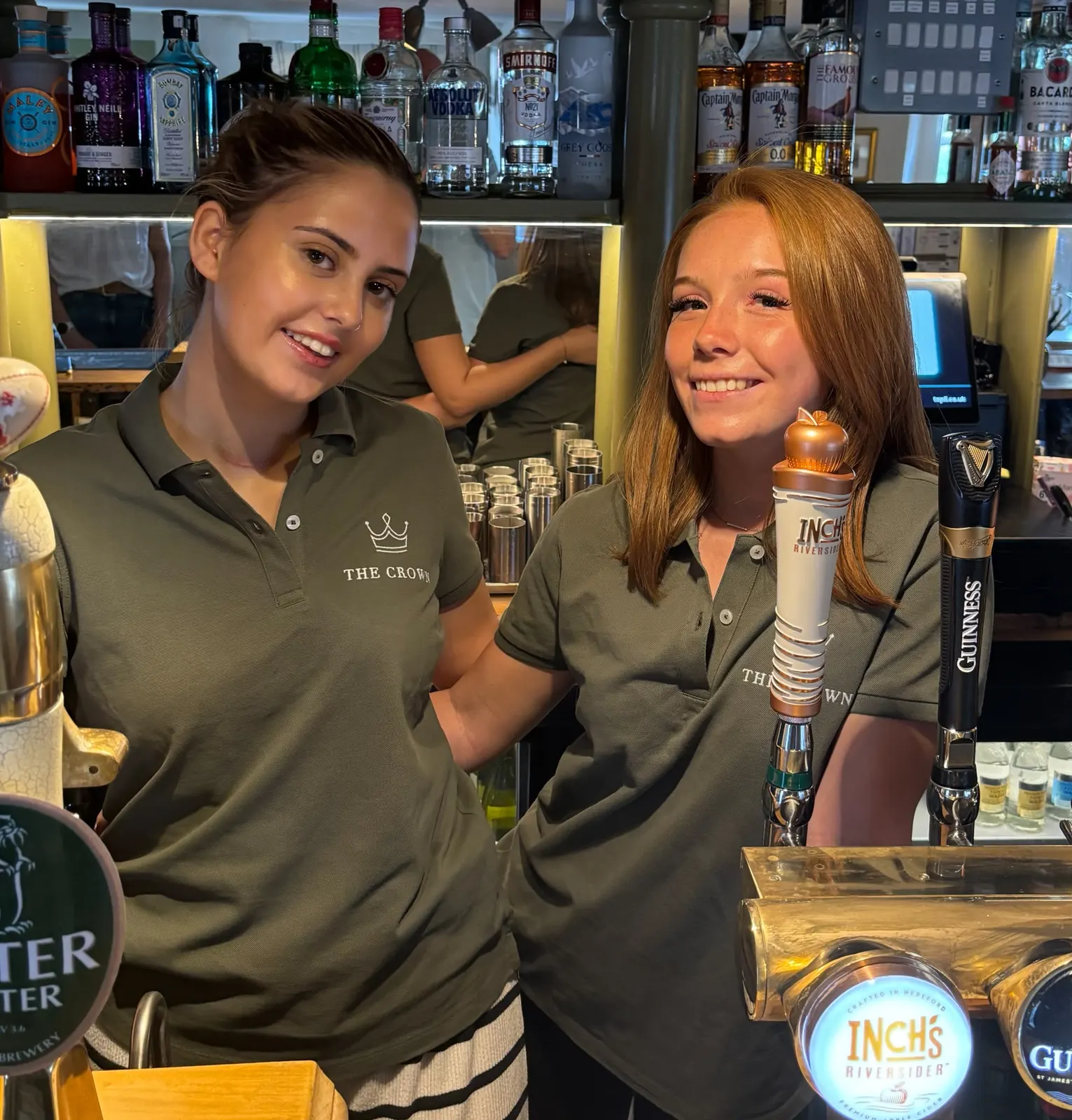 Eliza and Heather behind the bar at The Crown, in Crown polo shirts beside the beer taps